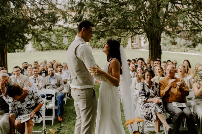 Picture showing outdoor wedding ceremony in Goldney Gardens. Couple are facing each other gazing into each other's eyes. The groom stands tall above the bride. Guests are sat on chairs, smiling and clapping